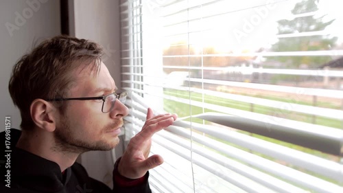 Man in glasses peeking through window blinds.