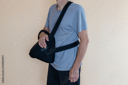An older adult man stands wearing a black arm sling to support his injured arm against a plain, light-colored wall.