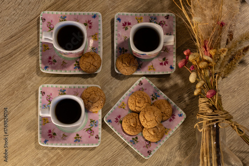 Four cups of coffee with cookies on floral saucers over a wooden table beside a bouquet of dried flowers, representing the social and cultural coffee tradition in Brazil.