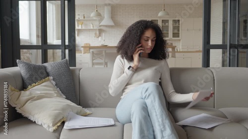 Worried Curly Woman Talking On Phone And Reviewing Financial Documents At Home