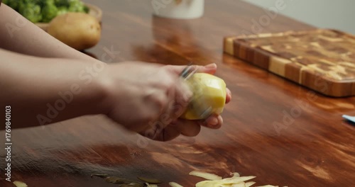 Close up of hands peeling fresh potato with peeler on wooden table, preparing ingredients for homemade vegan meal, cooking process, healthy food, rustic kitchen atmosphere, natural lifestyle