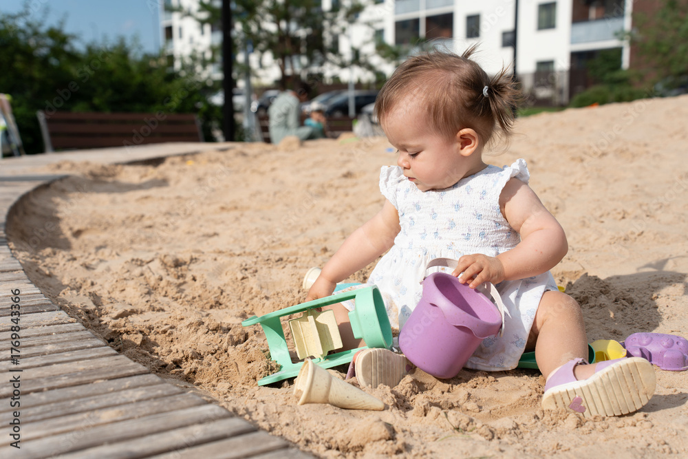 Naklejka premium a young child in a white dress sits on a sandy playground, engaged in play with sand toys, under sunlit conditions, surrounded by urban greenery and modern architecture, playful, build, explore