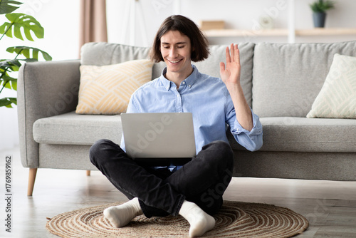 Foto In a bright living room, a young man sits cross-legged on the floor, smiling as he waves during a video call