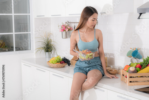 Beautiful Asian woman in blue athletic wear sits on a white kitchen counter, looking at a bowl of fresh salad. Surrounded by fruits and vegetables, she embodies a healthy lifestyle.