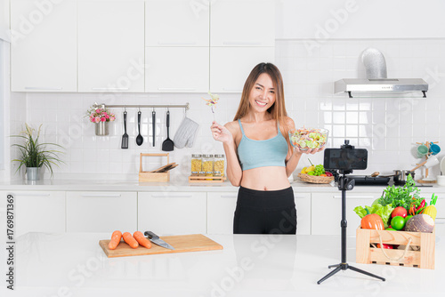 Smiling young woman in blue workout clothing prepare a healthy salad in her modern kitchen while recording a video on her smartphone, sharing her healthy lifestyle for online.