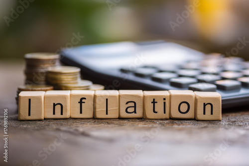 Wooden blocks spelling inflation with stacked coins and calculator in background symbolizing rising prices and financial analysis.