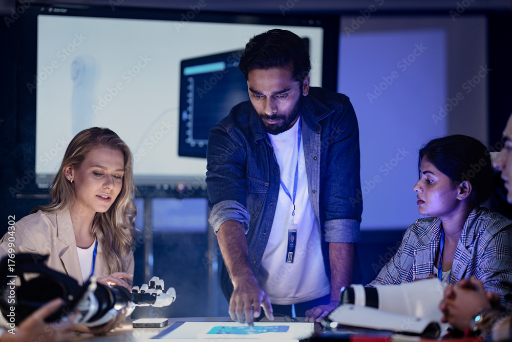 Fototapeta premium A robotics engineering team in an RD lab reviews a prototype. They are designing an AI powered robotic arm, analyzing data on a screen and a smart table.