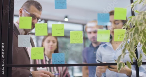 Work colleagues discussing startup project ideas. Team of caucasian business people standing near glass wall with sticky notes. Scrum methodology with colorful stickers. Low angle view focus on window
