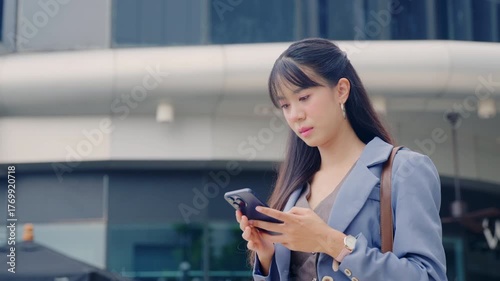 Young business businesswoman walking outdoor, focused on mobile phone while traveling, modern office buildings backdrop, urban professional woman navigating bustling city street environment.