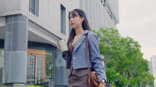 Young happy business businesswoman walking outdoor city street, holding travel mug, carrying shoulder bag, modern office building background, professional enjoying daily urban commute.