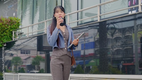 Young businesswoman walking outdoor, carrying coffee and laptop, modern office buildings framing urban city travel route, portraying successful business professional lifestyle.
