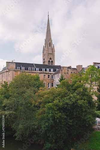 Beautiful cityscape of Bath, England, with historic stone buildings and a church spire overlooking the Avon River on a bright, partly cloudy day.