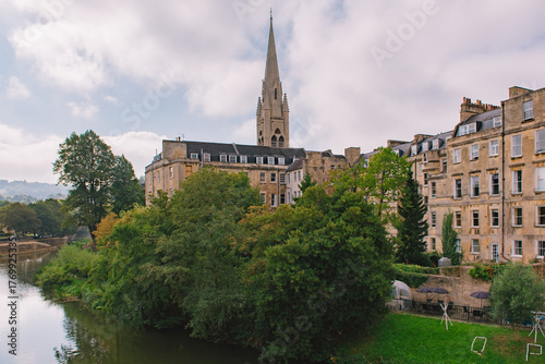 Beautiful cityscape of Bath, England, with historic stone buildings and a church spire overlooking the Avon River on a bright, partly cloudy day.