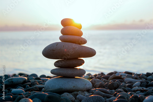 Balancing cairn of smooth stones on a seashore, illuminated by the soft rays of sunset, with the calm water surface in the background