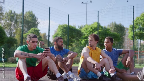 Attractive athletic diverse multi ethnic basketball players sitting on outdoor court, communicating and discussing match while friends taking a rest after streetball game in morning.