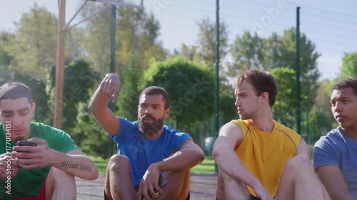 Portrait of handsome sporty fit diverse multiethnic male basketball players enjoying leisure, friendly chatting and sharing while sitting on outdoor court after streetball match at sunrise.
