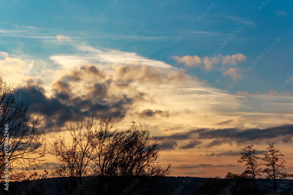 Fototapeta premium Stunning sunset sky ablaze with golden and orange hues, contrasted by dark, brooding clouds. Bare tree silhouettes frame this captivating, dramatic twilight scene