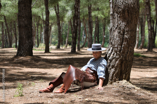 Schilderij op canvas A young, pretty woman dressed in riding breeches and riding clothes and a typical Spanish hat rests seated in the shade of a pine tree on a pilgrimage day in Spain