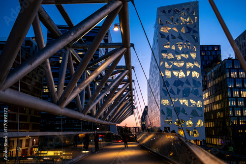 Panoramic view of the Akrobaten bridge in Oslo showing the Barcode architecture and skyline illuminated by warm evening light, modern urban structures and dynamic reflections