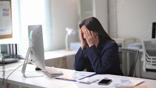 A woman is sitting at a desk in a professional setting, typing on a computer keyboard. She is wearing a blue suit and she is focused on her work. The room is well-lit and organized, with a few books