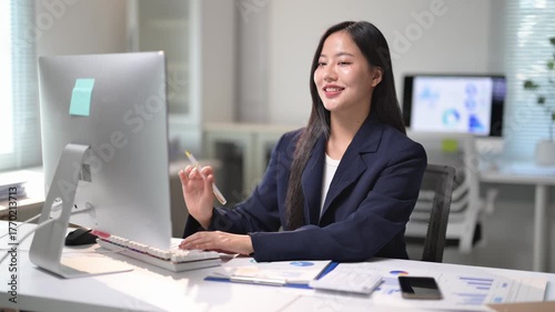 A woman in a business suit is sitting at a desk with a computer monitor in front of her. She is holding a pen and she is typing or writing something. Concept of professionalism and focus