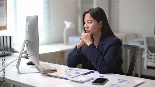 A woman is typing on a computer in a professional setting. She is wearing a suit and she is focused on her work. The room is well-lit and has a desk with a computer, a keyboard, and a mouse