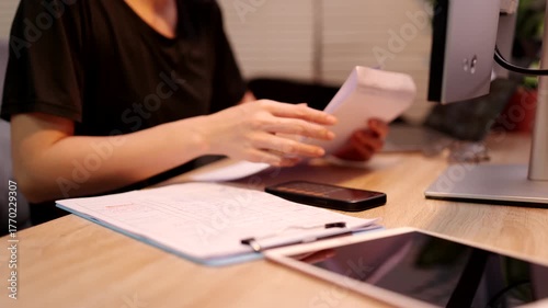A person reviews tax documents and calculates financial data using a smartphone and computer at a desk.
