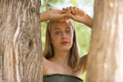 Portrait of a young woman, blonde, blue eyes, slim, wearing a green dress, looking sweetly and tenderly at camera, leaning against tree trunks. Concept: femininity, innocence, youth.
