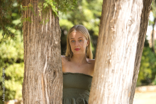 Portrait of a young woman, blonde, blue eyes, slim, wearing a green dress, looking sweetly and tenderly at camera, leaning against tree trunks. Concept: femininity, innocence, youth.