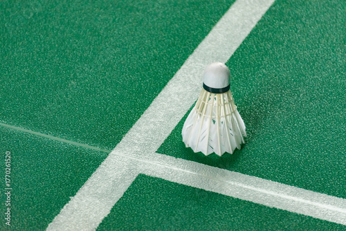 A solitary shuttlecock is positioned on the corner where two white court lines meet on a bright, aqua-green textured floor, symbolizing the corner of the service area.
