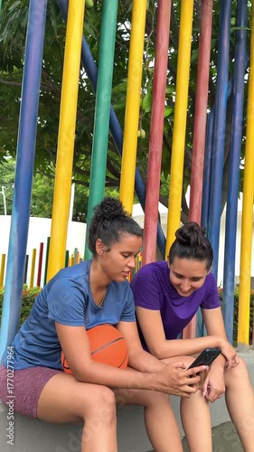 Two smiling female basketball players sitting together in a park watching a smartphone