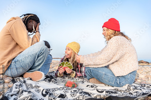 Group of diverse happy friends playing cards and enjoying a picnic on a rocky beach