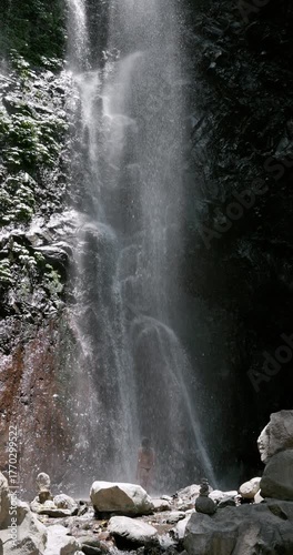 Woman in swimsuit staying under falling water on waterfall.