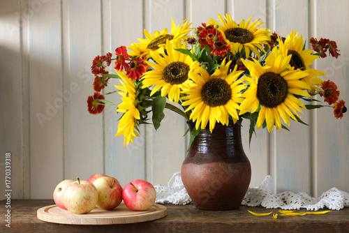 A bouquet of sunflowers in a clay jug and rustic apples are placed on a cutting board. summer still life with garden flowers and fruits.