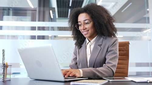 Smiling african american businesswoman typing on laptop while sitting at desk at workplace in modern business office. Black woman employee works on a computer, is engaged in a project, busy with tasks