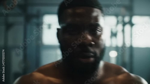 Extreme close-up video of a powerful black man's sweating forehead, tracking back to his determined face in a dark gym with dramatic rim lighting. Concept of unyielding will