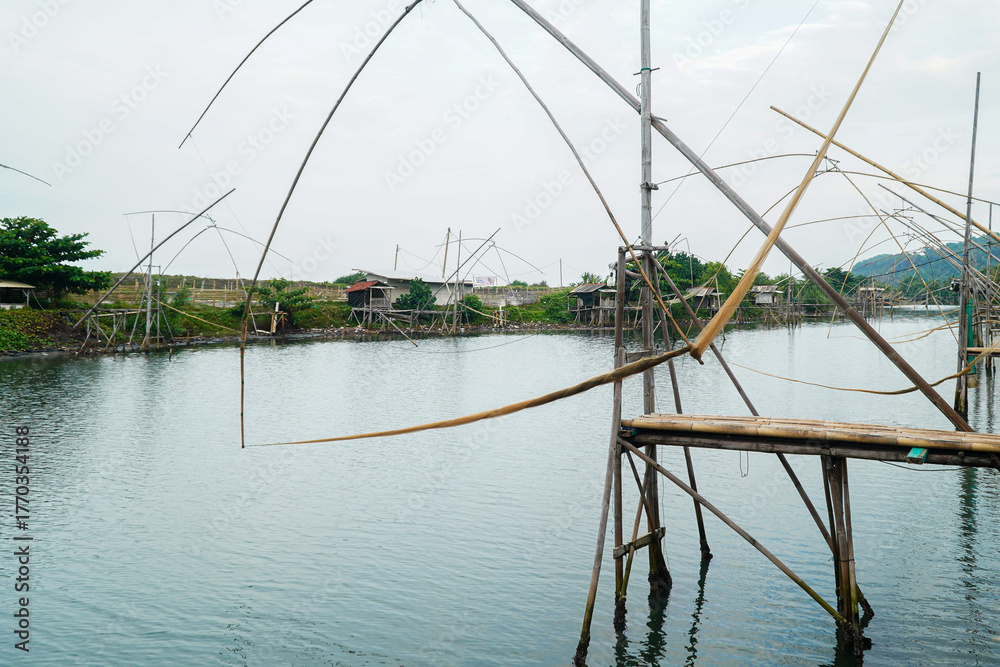 Fototapeta premium A tranquil scene featuring traditional fishing structures standing tall in calm water, embodying a serene and timeless fishing tradition, with the still water mirroring the overcast sky.