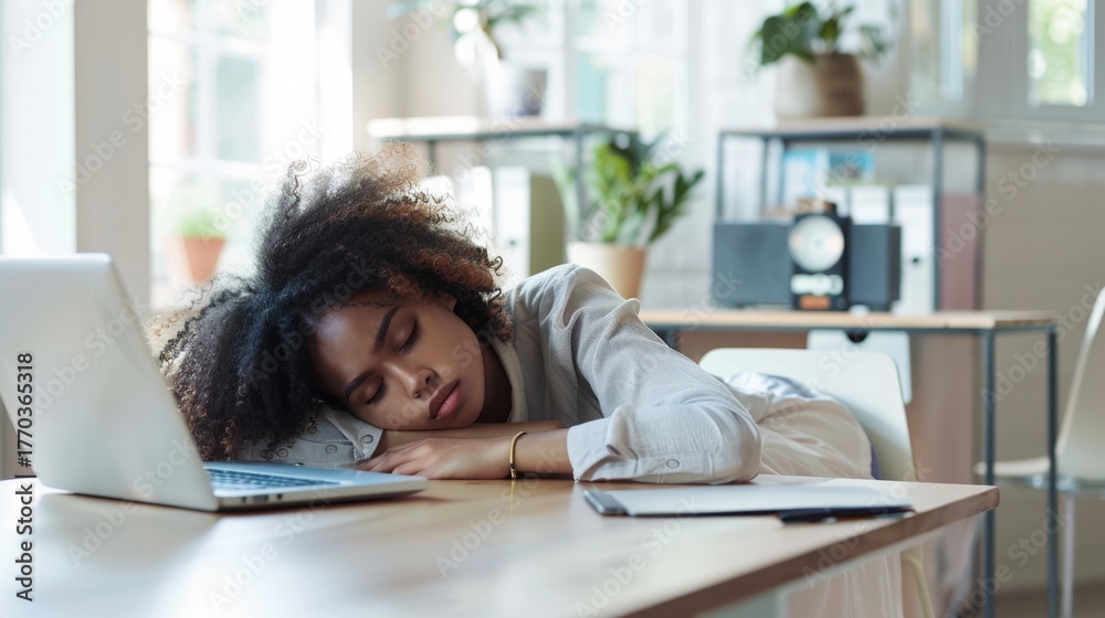 Obraz premium Young African woman with curly hair resting on a desk in a modern office. A laptop and notebook are visible on the table. Indoor setting with plants in the background.