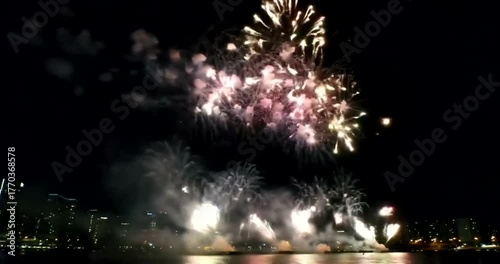 Spectacular fireworks display explodes in the night sky over a city skyline reflected in the water.