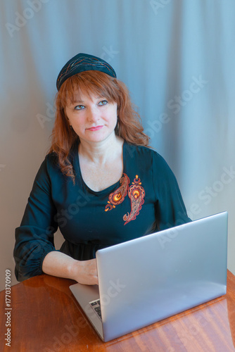 A beautiful young woman is working or playing or shopping on a laptop in a stylish modern dress and a hair band in the shape of the Russian national kokoshnik headdress