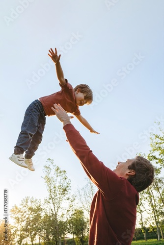 Joyful father playing with his smiling young son, tossing up and catching him in a park at sunset. Family bonding, happy childhood and Fathers day concept. Copy space