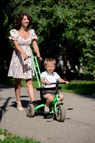 A cute baby sits on a tricycle. The child goes for a walk with his mother in the park. A happy smiling boy.