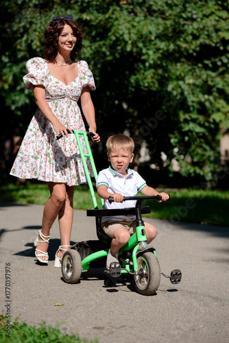 A cute baby sits on a tricycle. The child goes for a walk with his mother in the park. A happy smiling boy.