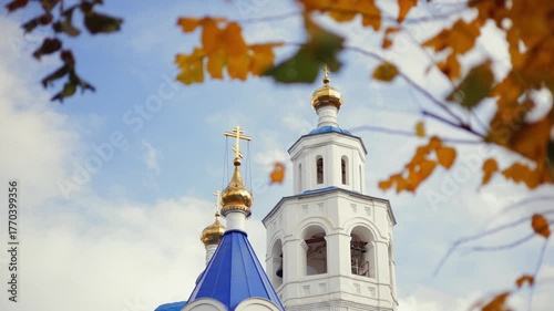 Golden leaves frame blue domed orthodoxy church towers against cloudy sky, yellow autumn leaves in foreground.