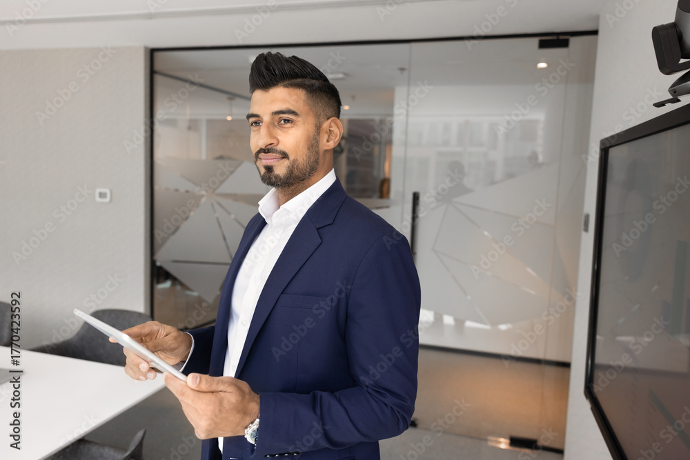 Fototapeta premium Positive pensive handsome young Indian business professional man holding tablet