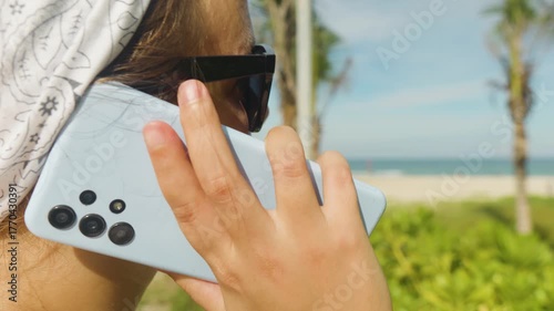 Woman making a phone call on a light blue mobile phone, wearing a bandana and sunglasses, while enjoying a summer vacation day at the beach with palm trees and ocean in the background