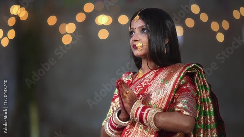 A newly married woman dressed in a red saree with traditional jewelry stands with folded hands in prayer. The soft light and bokeh background create a peaceful and elegant scene.