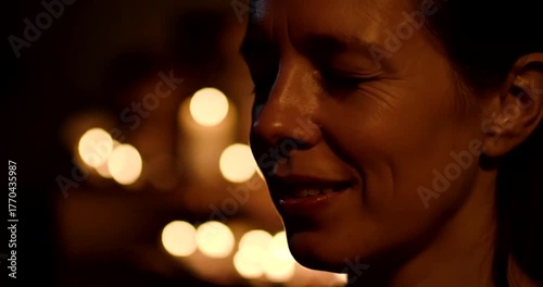 Woman smiling gently with her eyes closed in a dark room illuminated by warm candlelight.