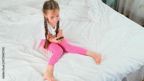 Smiling Caucasian child girl sitting on the bed with tv remote