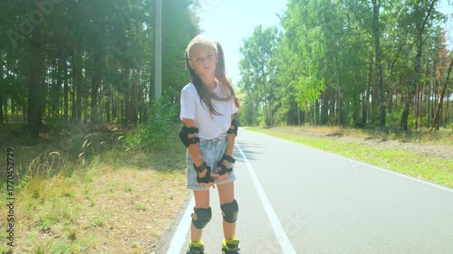 Child girl in casual clothes roller skating on road in forest on sunny summer day.
